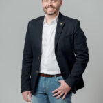 Homme souriant en blazer noir et chemise blanche devant fond gris neutre