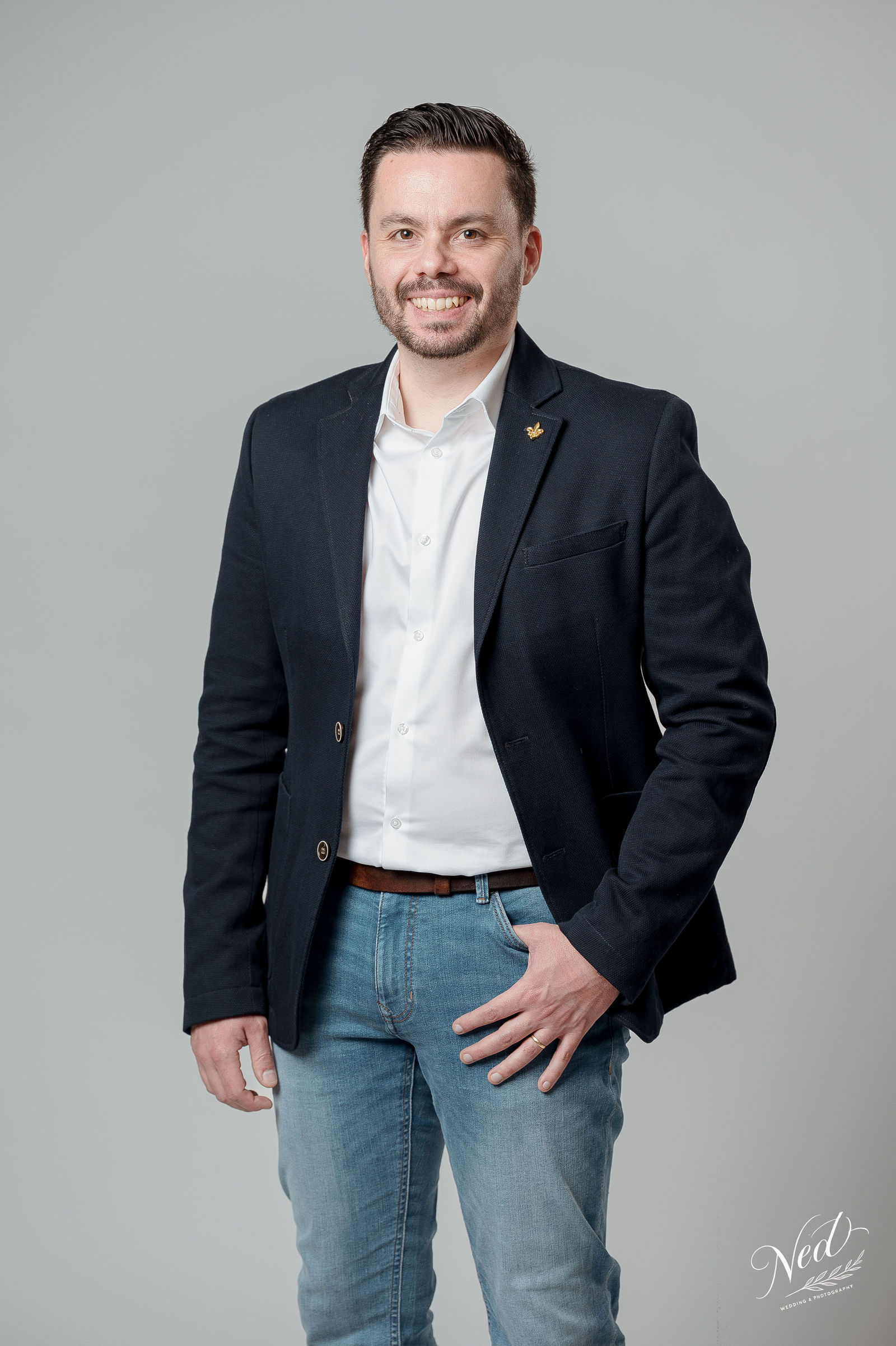 Homme souriant en blazer noir et chemise blanche devant fond gris neutre