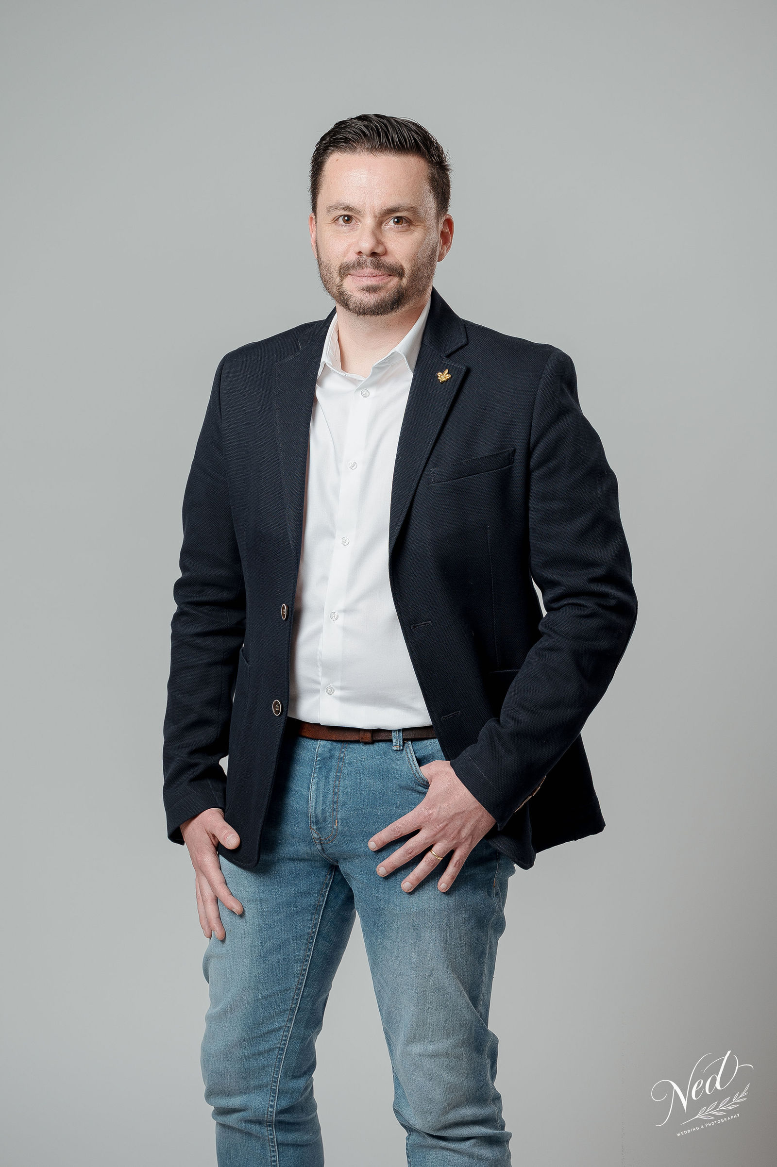 Homme en blazer noir et chemise blanche pose face au photographe, main dans la poche, fond gris neutre