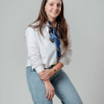 Femme souriante en chemise blanche et foulard bleu posée devant fond neutre gris