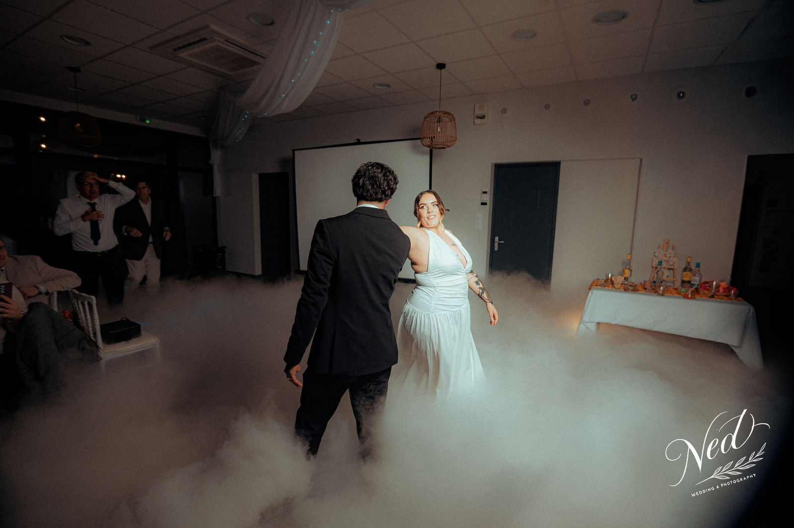 Mariée en robe blanche et marié en costume noir dansent ensemble sur la piste de danse avec effet brouillard, invités en arrière-plan