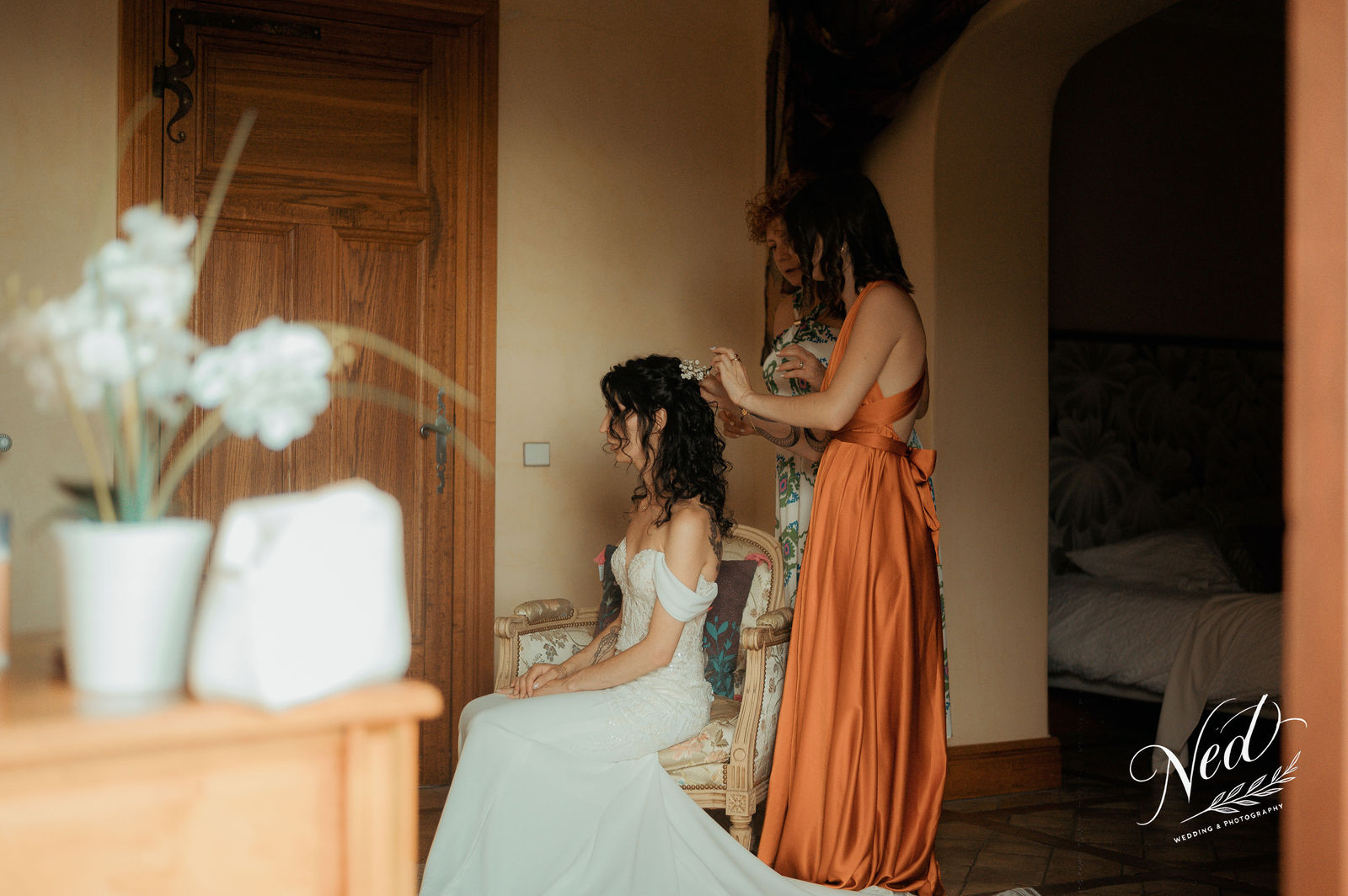 Demoiselle d'honneur en robe orange coiffant la mariée en robe blanche assise sur une chaise dorée, fleurs blanches au premier plan