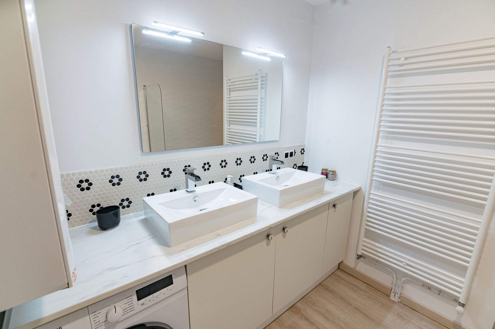 Salle de bain épurée avec deux vasques blanches, miroir éclairé, carrelage géométrique noir et blanc, meubles crème