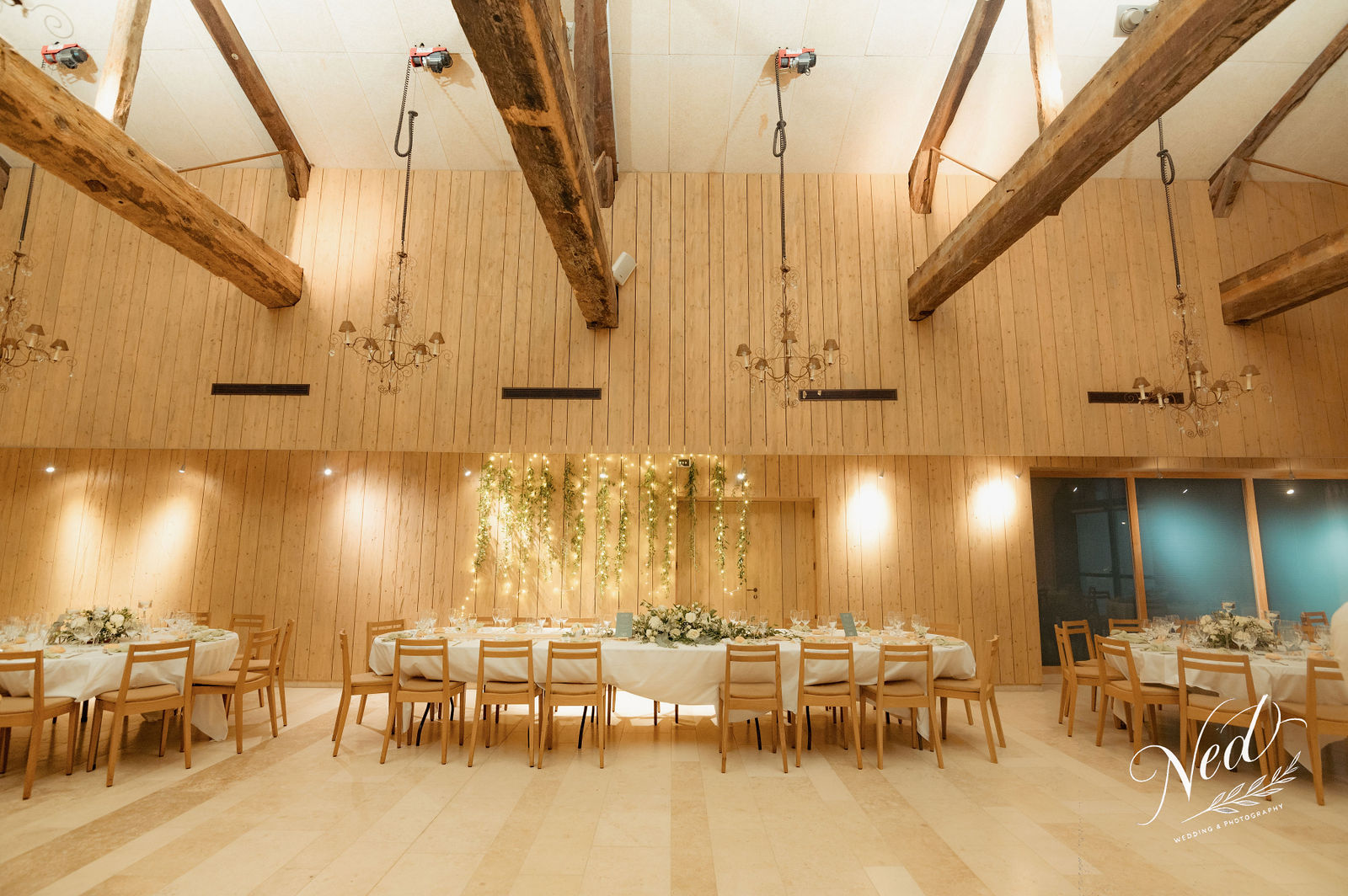 Salle de mariage en bois clair avec poutres apparentes, tables rondes nappe blanche, guirlandes lumineuses et fleurs suspendues