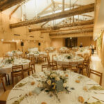 Salle de mariage avec poutres apparentes, tables rondes dressées avec fleurs blanches et or, chaises en bois clair, lumière naturelle par verrière