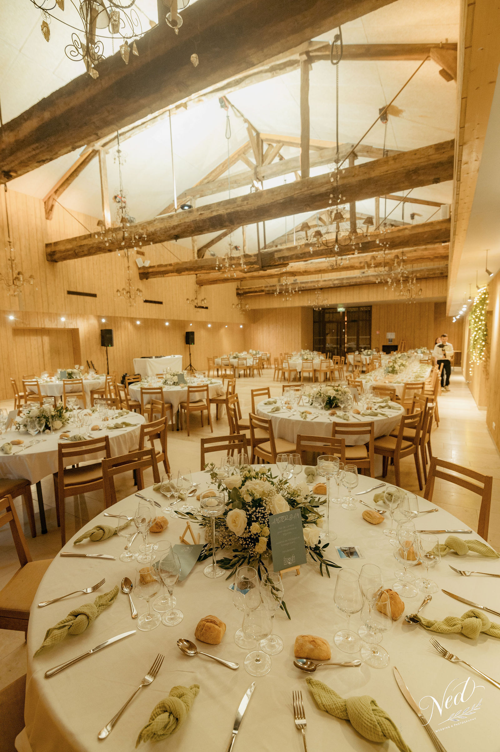 Salle de mariage avec poutres apparentes, tables rondes dressées avec fleurs blanches et or, chaises en bois clair, lumière naturelle par verrière