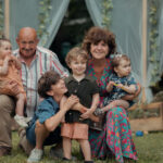 Grands-parents et quatre enfants posent souriants devant une maison avec volets bleus, assis et debout sur l'herbe