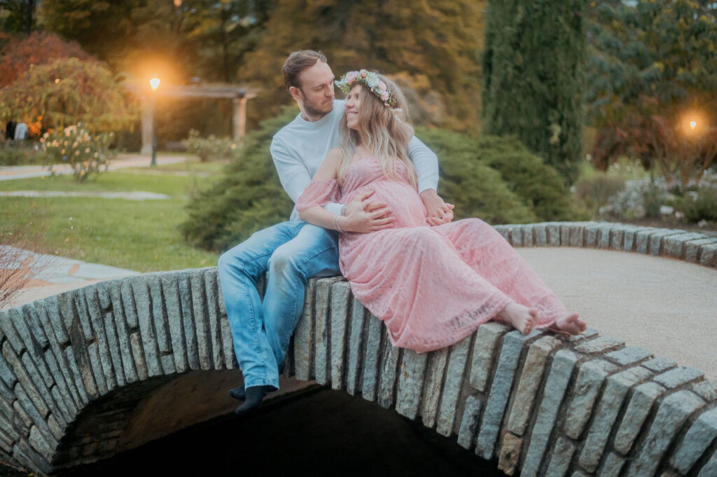 Femme enceinte en robe rose avec couronne de fleurs assise sur un muret en pierre, son compagnon derrière elle dans un parc en fin d'après-midi