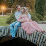 Femme enceinte en robe rose avec couronne de fleurs, assise contre un homme en pull blanc qui pose sa main sur son ventre
