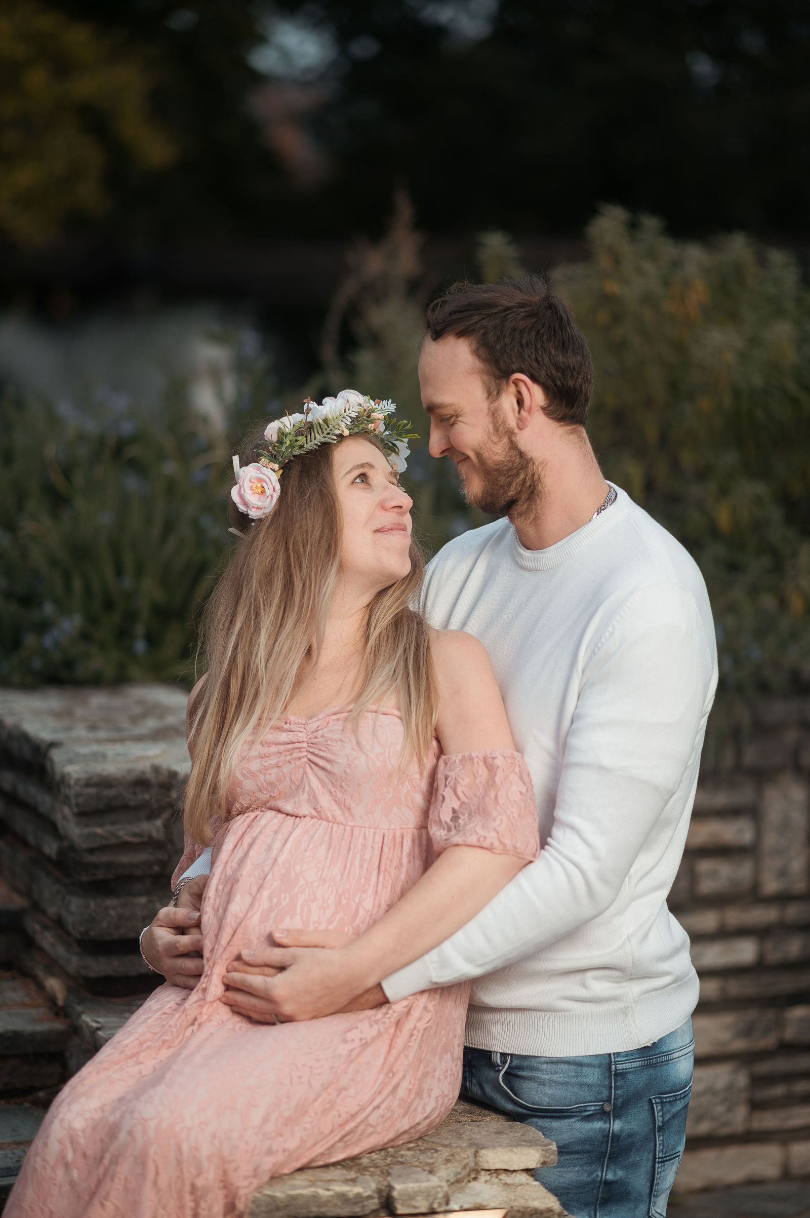 Femme enceinte en robe rose avec couronne de fleurs, assise contre un homme en pull blanc qui pose sa main sur son ventre