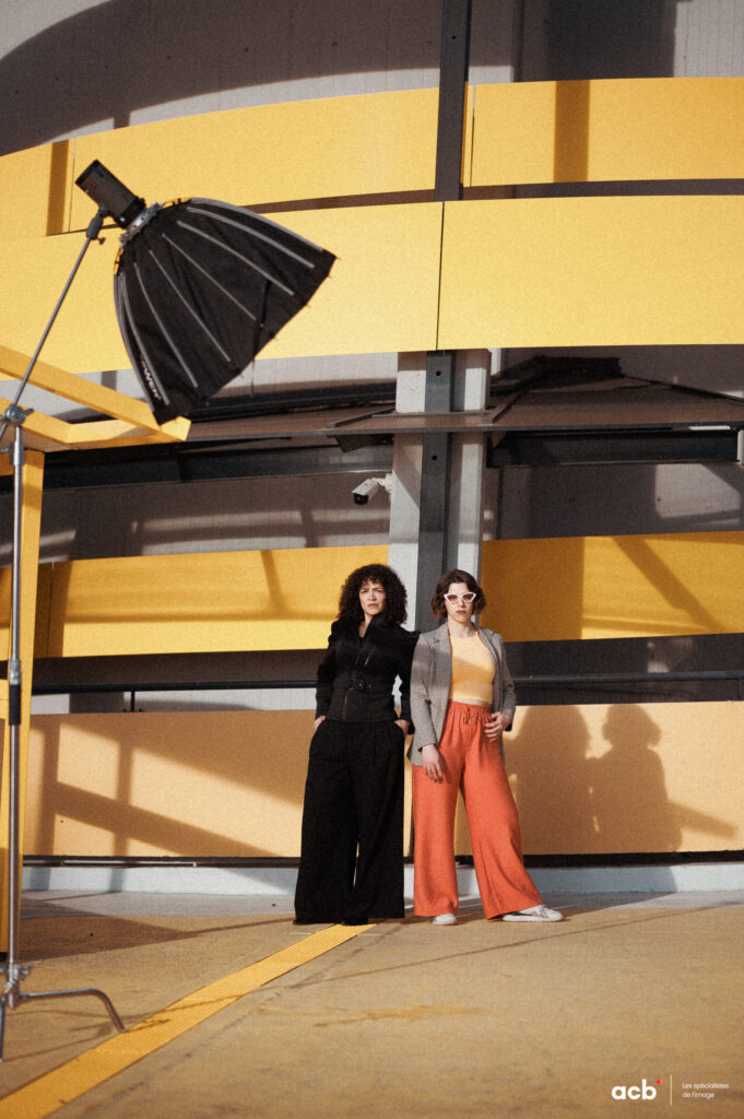 Deux femmes posent devant un mur jaune et noir en studio, softbox noir suspendu au-dessus, lumière directe créant des ombres nettes