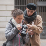 Formateur en bonnet noir et pull beige guide une participante qui tient un appareil photo reflex, en extérieur devant une facade grise.