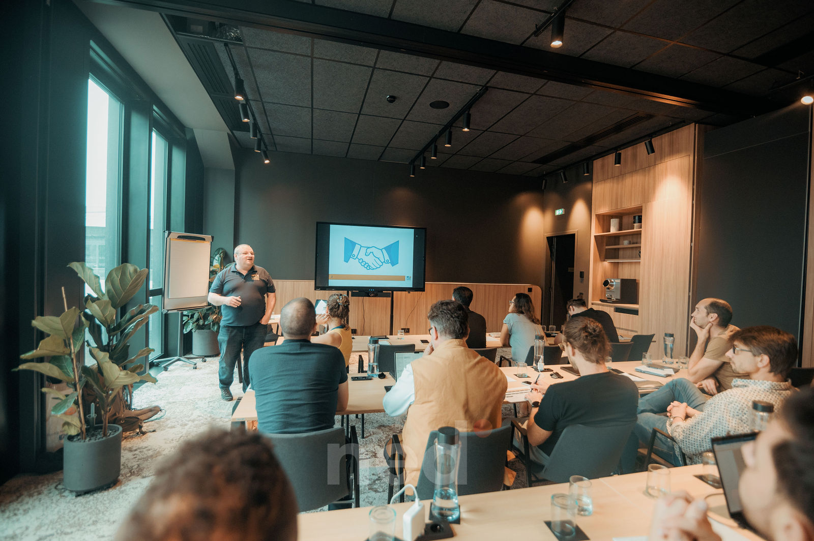 Formateur en polo bleu présente devant dix participants assis autour de tables dans une salle moderne, écran de projection visible en arrière-plan