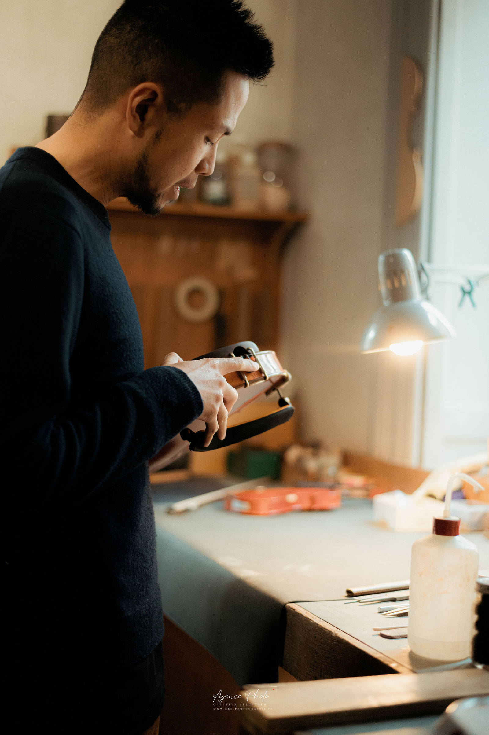 Homme en pull noir regarde son appareil photo sur un plan de travail, lumiere naturelle de cote eclaire son visage et le materiel photo