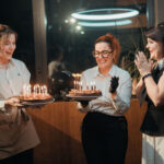 Trois femmes souriantes tiennent des gâteaux avec bougies allumées lors d'une fête en intérieur, lumière tamisée et néon bleu en arrière-plan