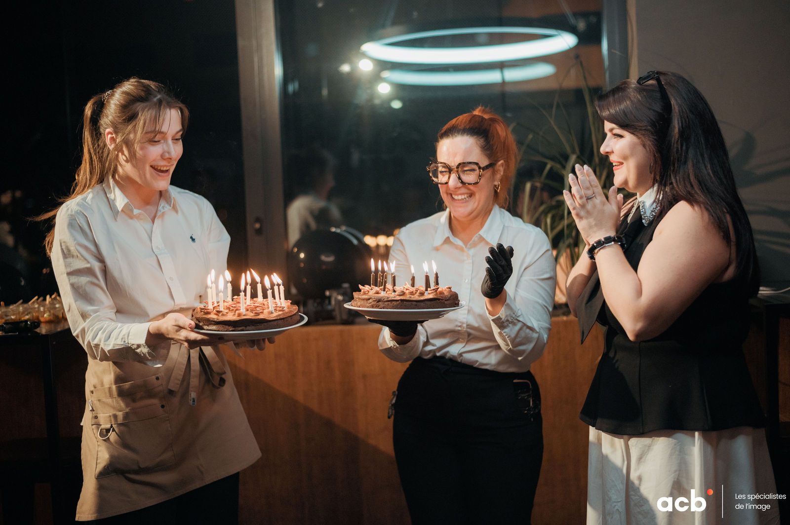 Trois femmes souriantes tiennent des gâteaux avec bougies allumées lors d'une fête en intérieur, lumière tamisée et néon bleu en arrière-plan