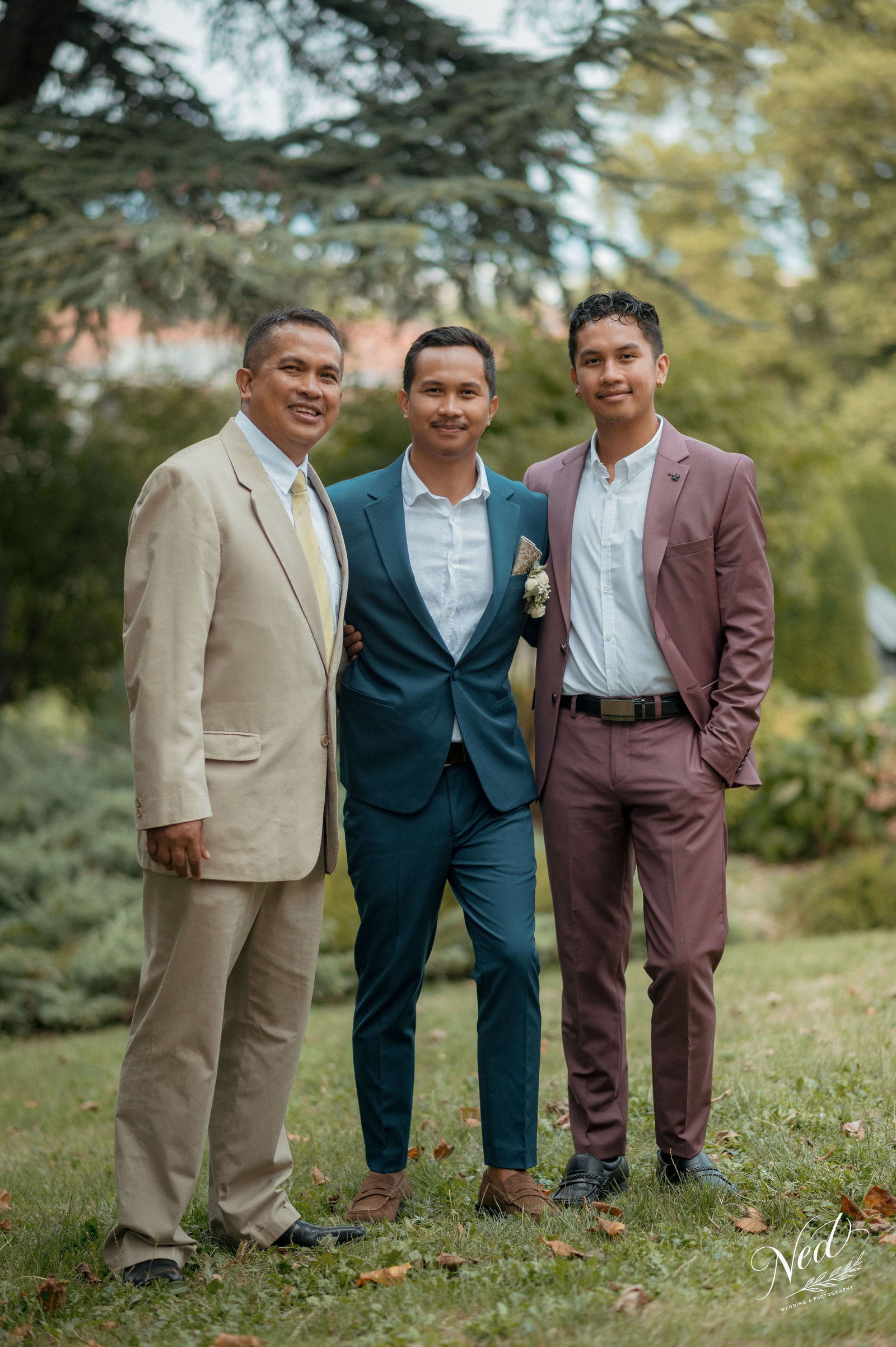Trois hommes souriants en costumes colorés posent ensemble sur l'herbe devant des arbres