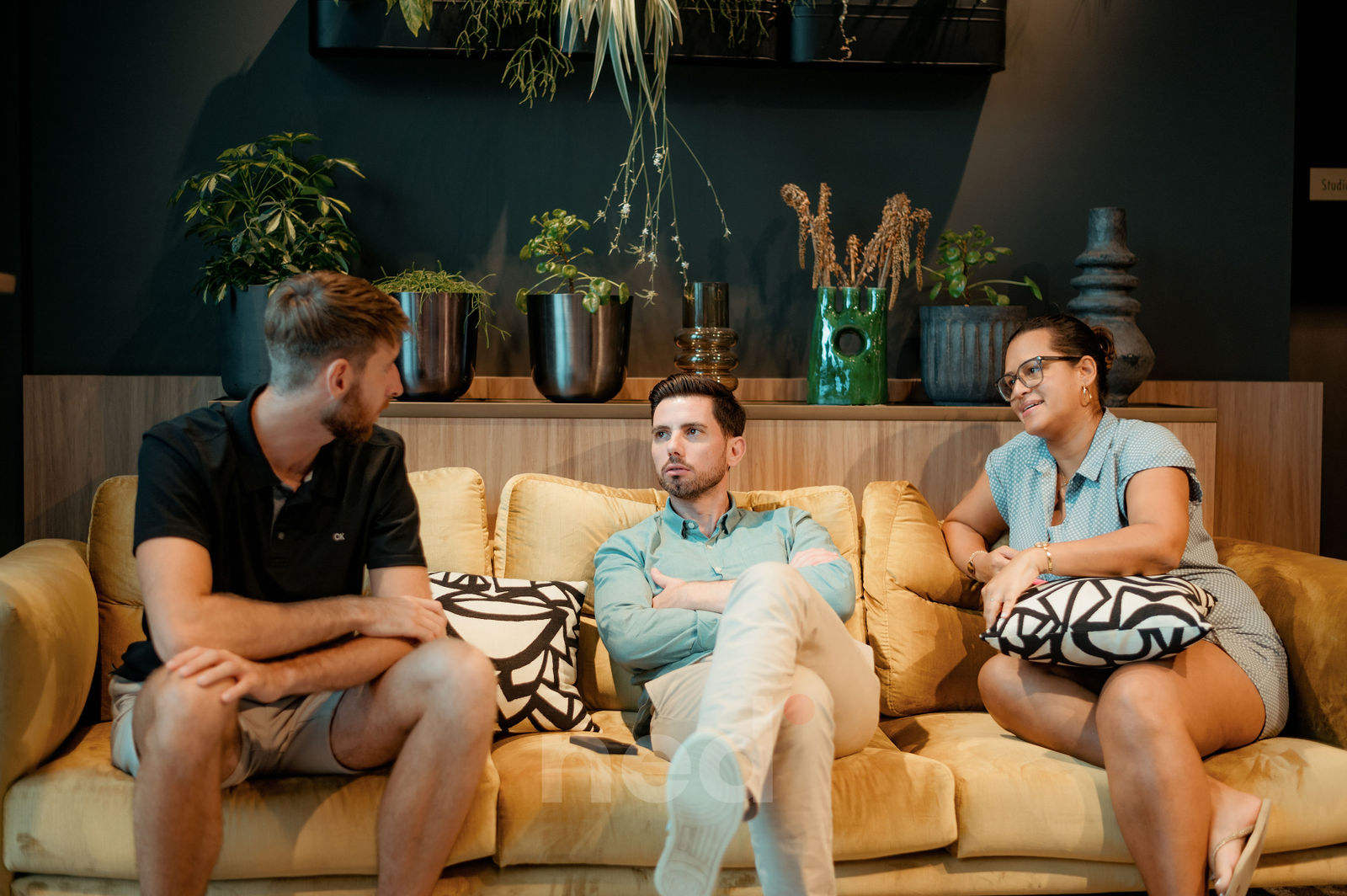Trois personnes assises sur un canapé beige dans un salon moderne, plantes vertes en arrière-plan sur étagère bois.
