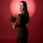 Femme en robe noire épaulée dénudée tenant un bouquet de fleurs rouges, fond rouge vif en studio