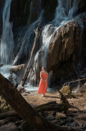 Femme en robe rouge corail debout face a une cascade, entouree de rochers moussus et bois mort en montagne