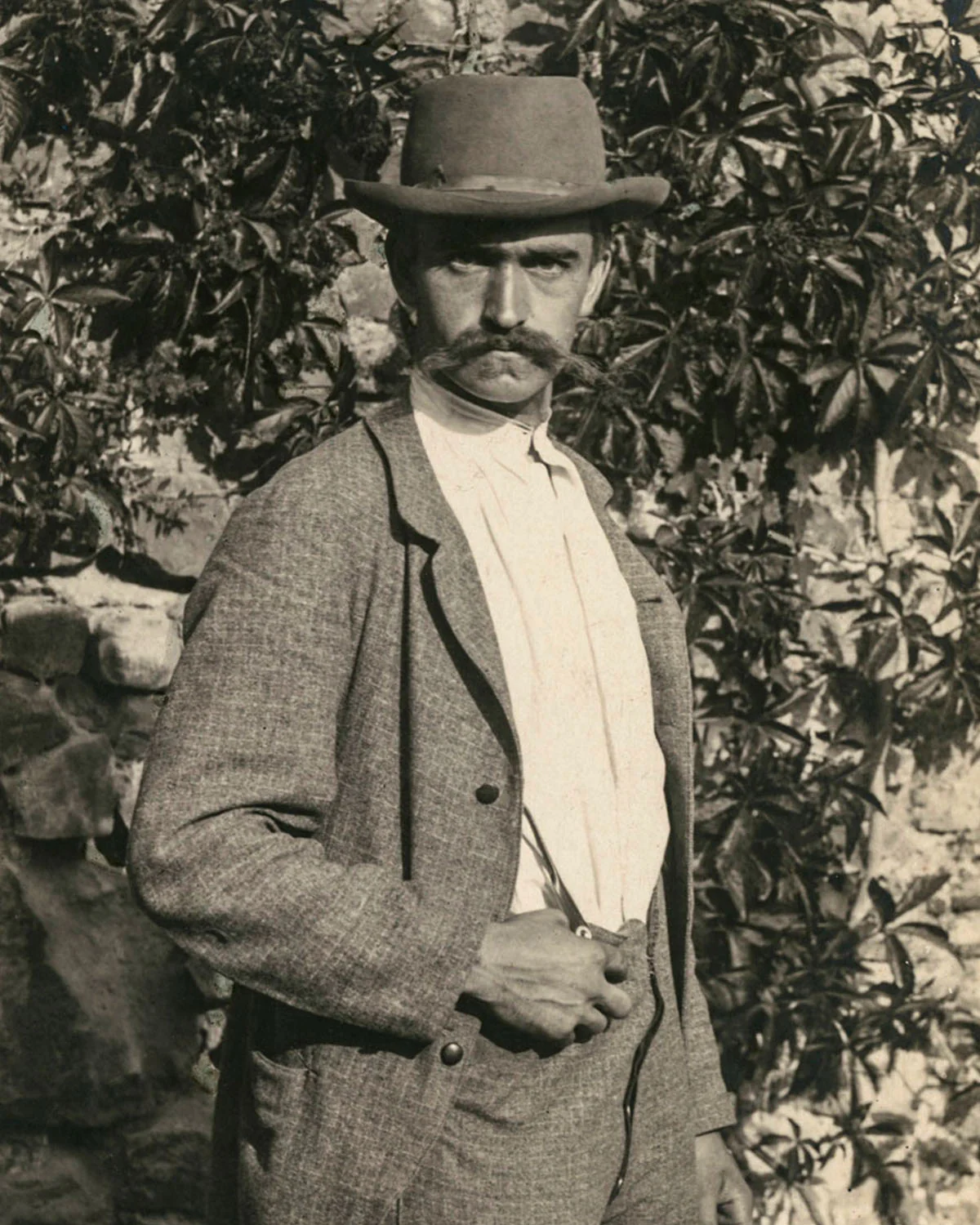 Homme en costume gris et chapeau posé devant un mur de feuillage dense, photographie en noir et blanc d'époque