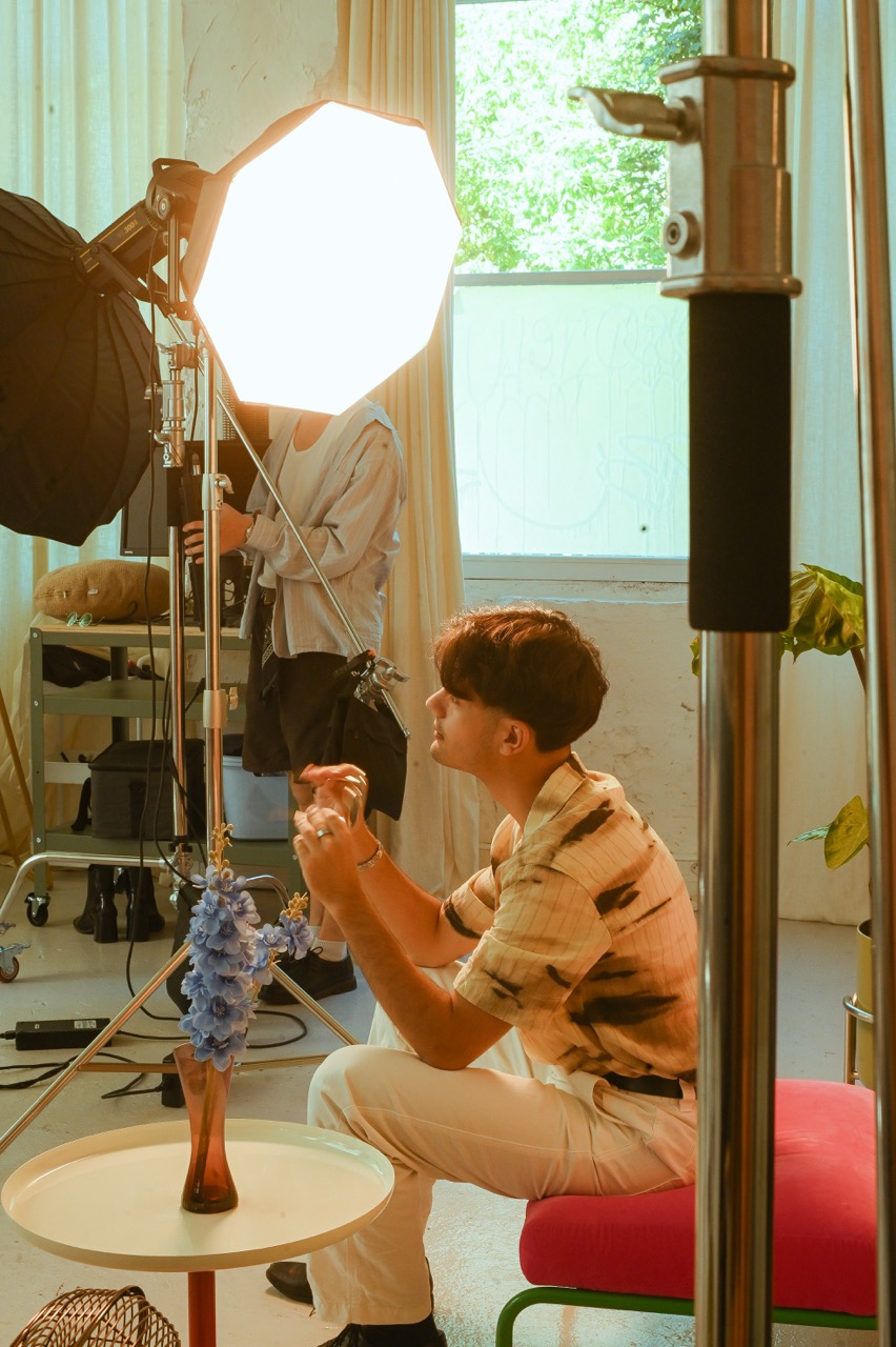 Photographe en chemise orange ajuste un projecteur LED octogonal face à un vase de fleurs bleues en intérieur, fenêtre verte en arrière-plan
