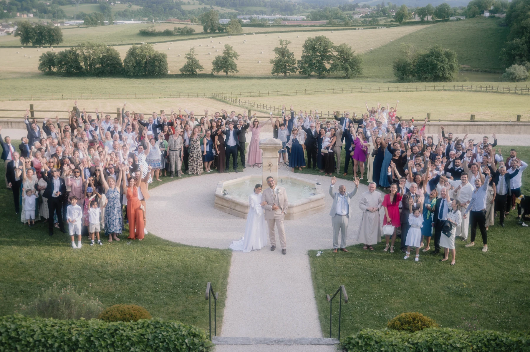 Photo de groupe du mariage : les mariés au centre près de la fontaine, les invités autour sur la pelouse