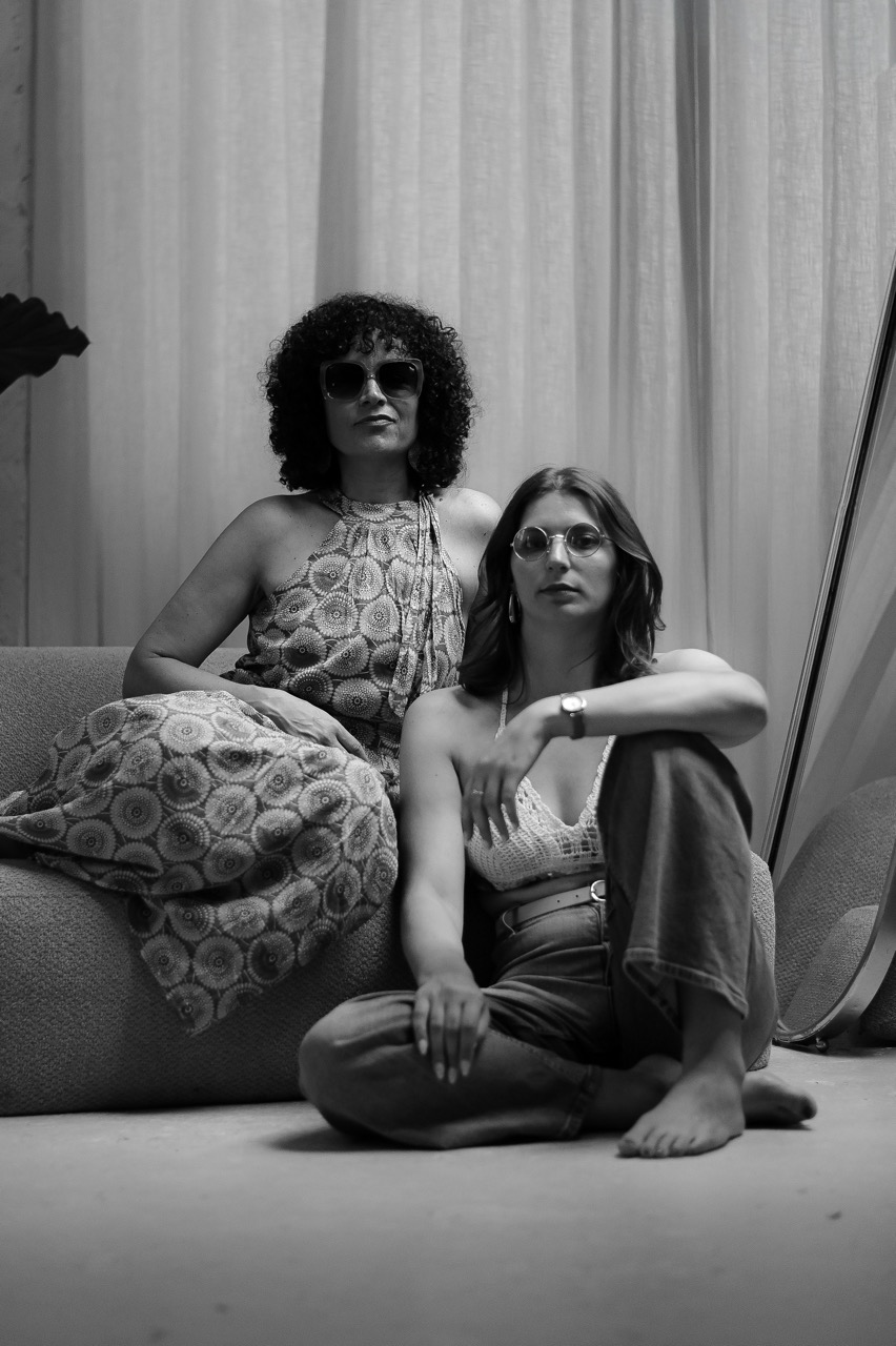 Deux femmes portant des lunettes de soleil posent sur un canapé devant des rideaux blancs en studio