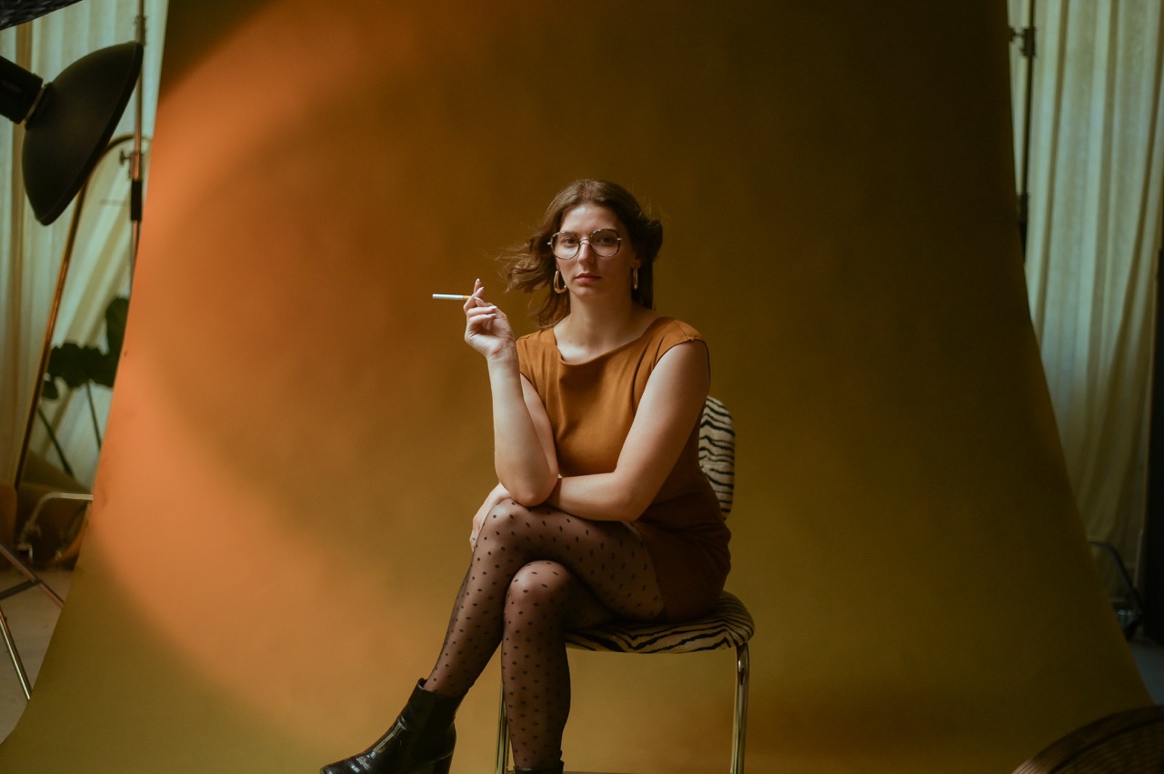 Femme assise sur une chaise portant des lunettes et un chemisier ocre, cigarette à la main, devant un fond orange uni en studio