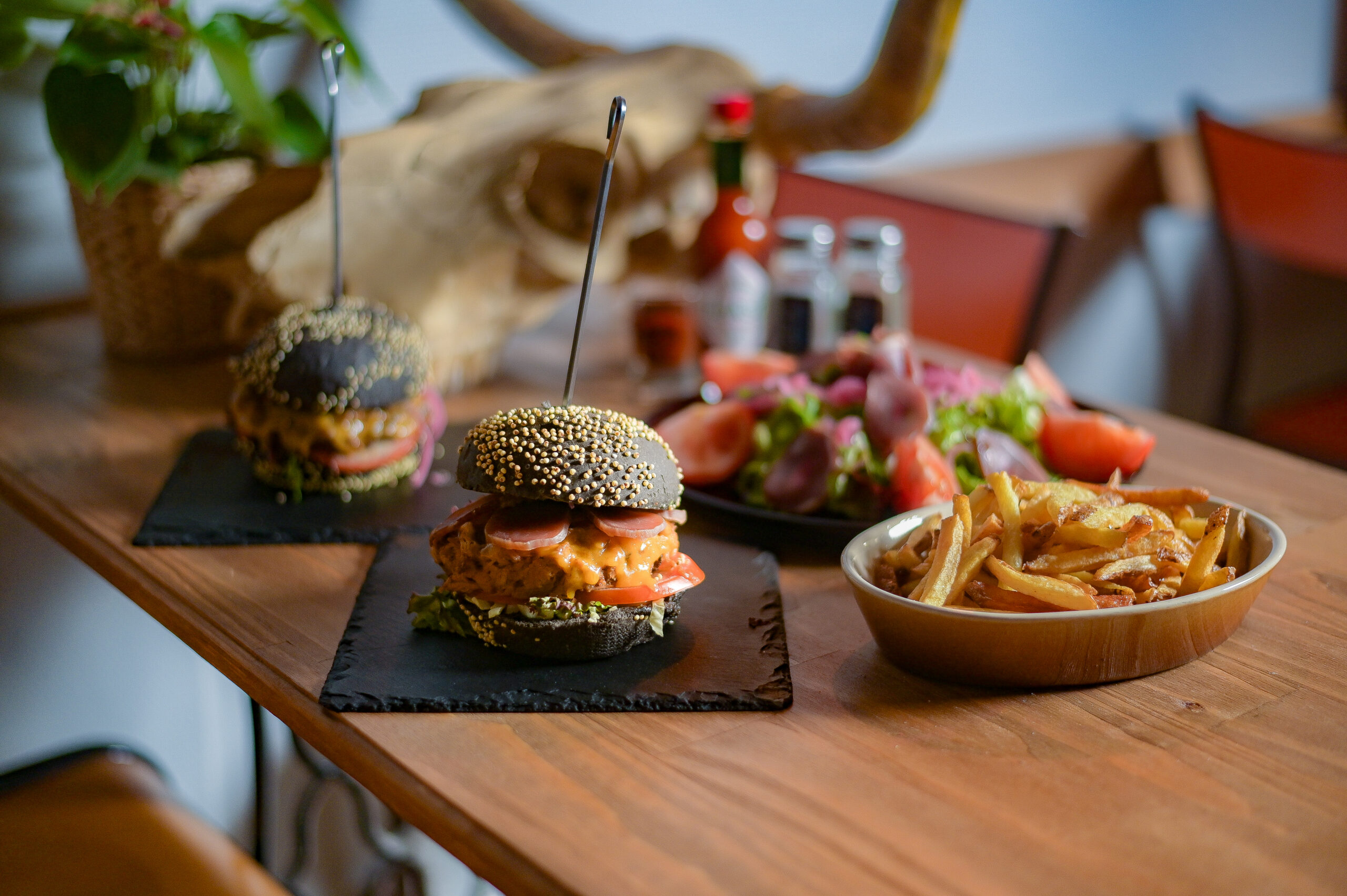 Deux burgers aux graines dorées sur ardoise noire avec frites en bol, table en bois clair, decor restaurant
