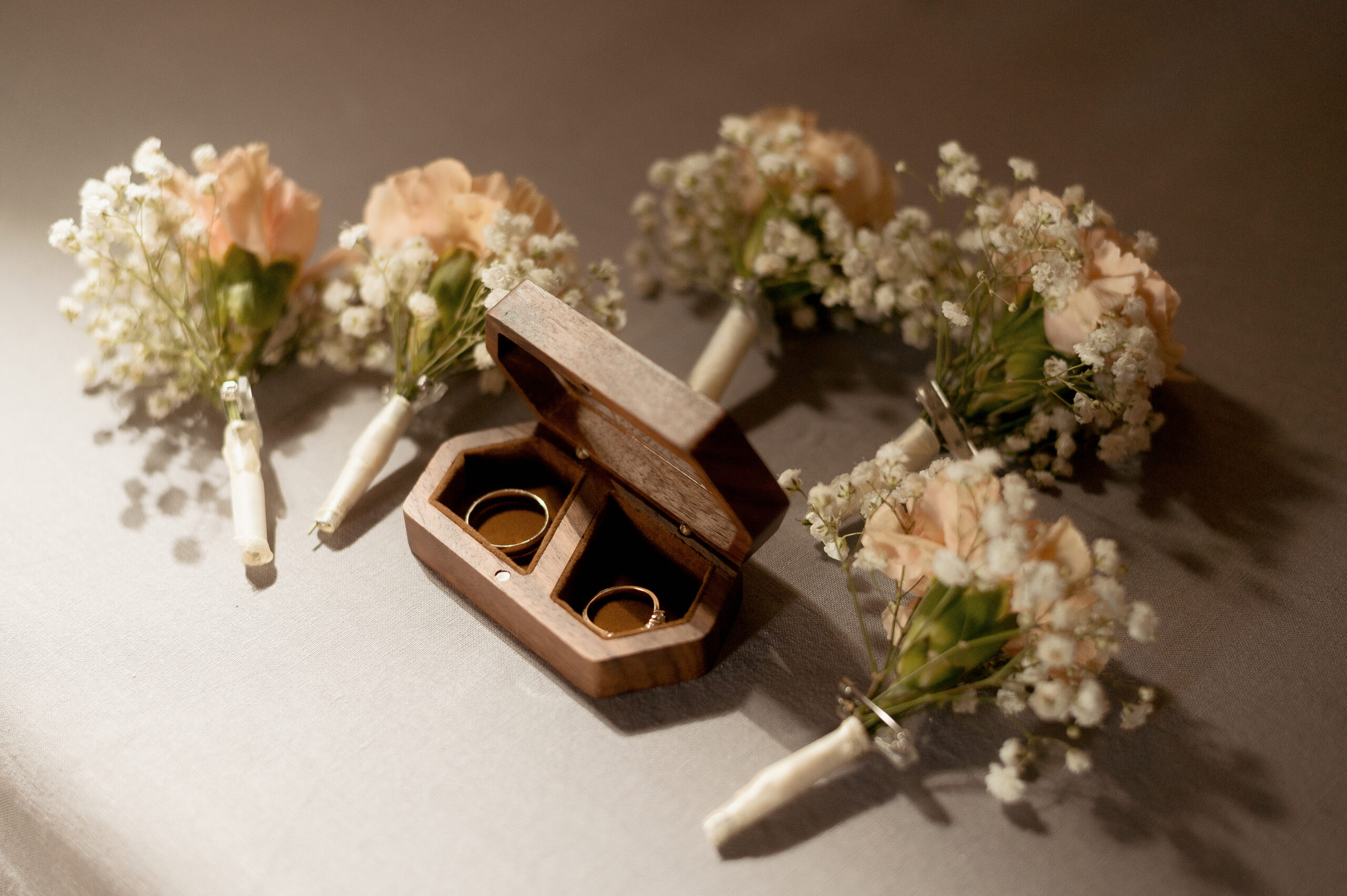 Deux alliances dorées dans un écrin en bois ouvert, entouré de petits bouquets de gypsophile blanche et fleurs pêche sur fond neutre