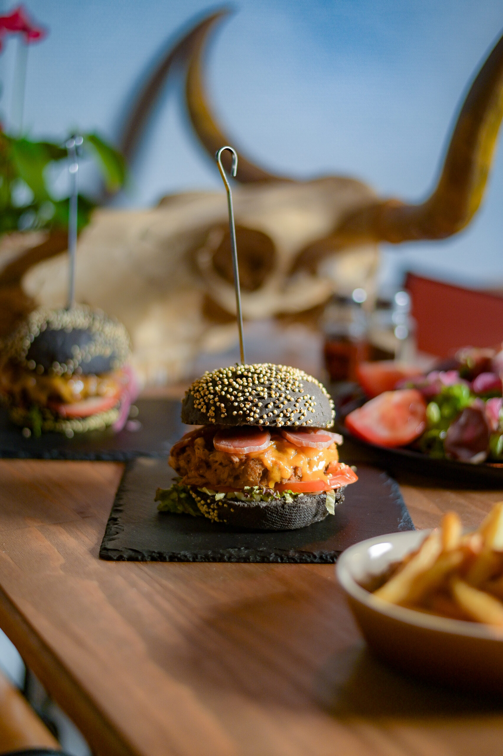 Petit burger au pain noir garni de saumon et légumes sur planche en ardoise, cornes de taureau en arrière-plan