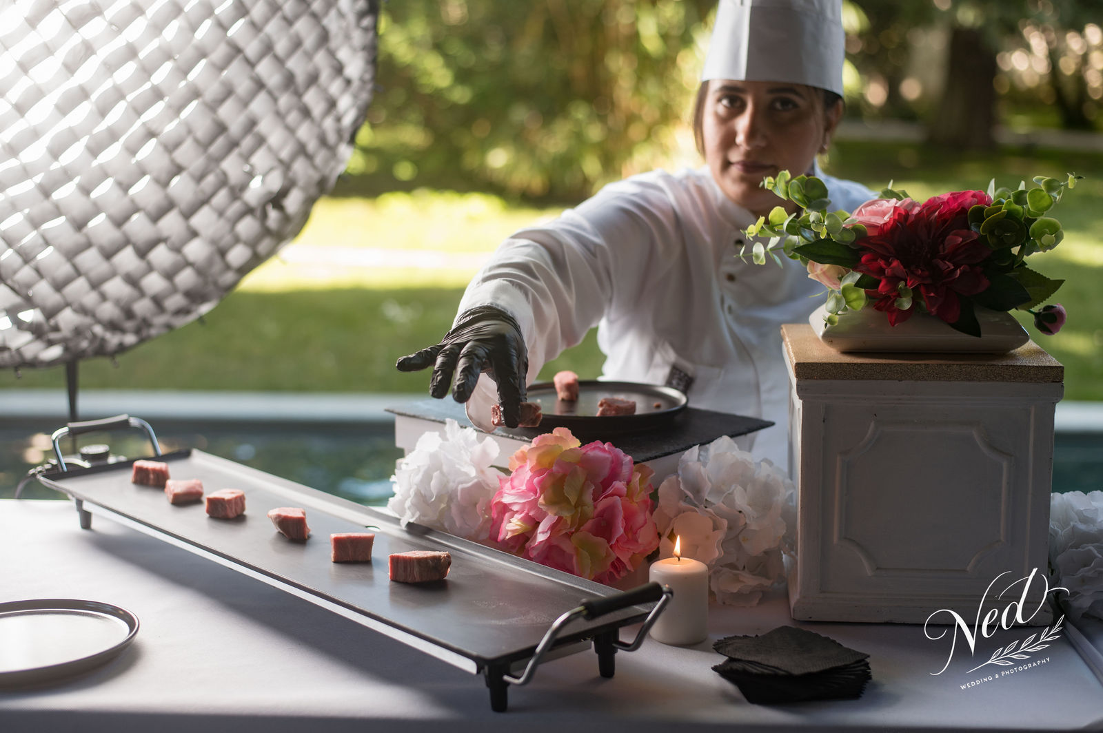 Chef en toque blanche dresse une assiette avec des cubes de viande devant des fleurs roses, jardin verdoyant en arrière-plan