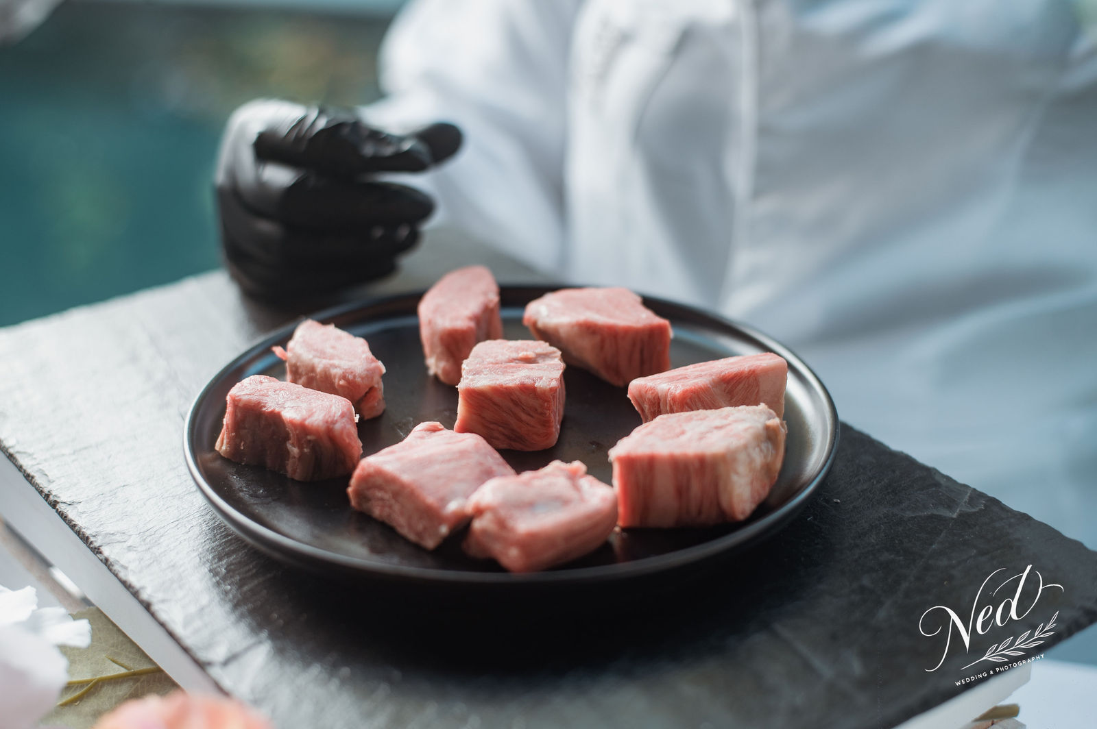 Chef en tenue blanche tenant une assiette noire avec des cubes de viande crue sur ardoise grise, fond bleu