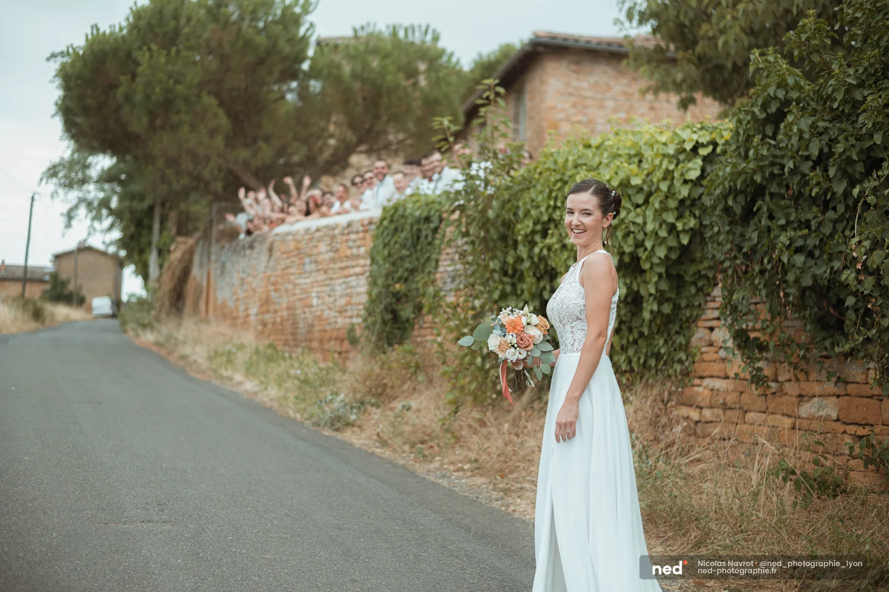 La mariée souriante marche vers le first look pendant que les invités observent depuis le muret en pierres dorées, Anse