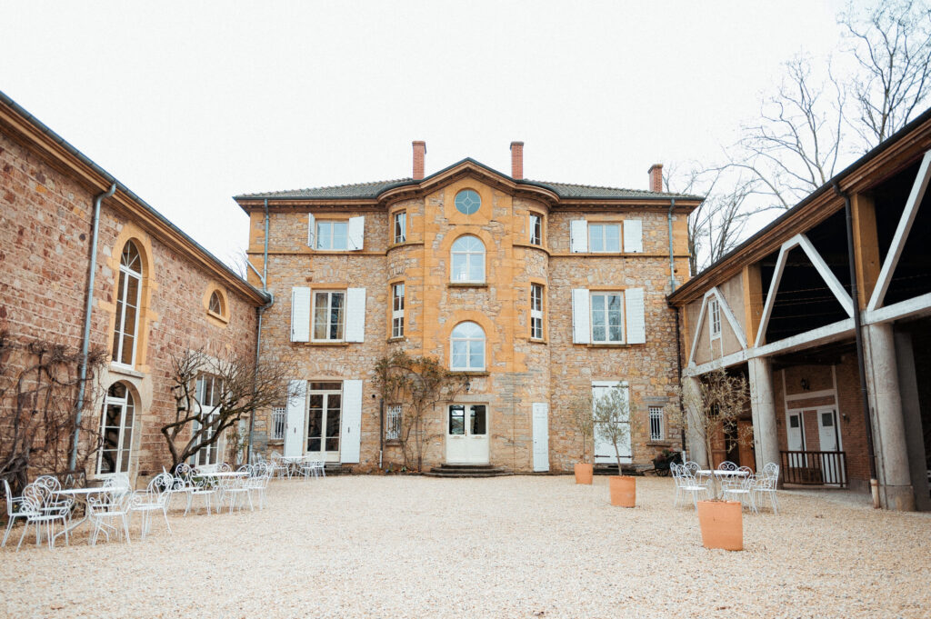 Cour interieure du domaine de Tourieux avec chaises blanches, batisse en pierre blonde et ciel nuageux