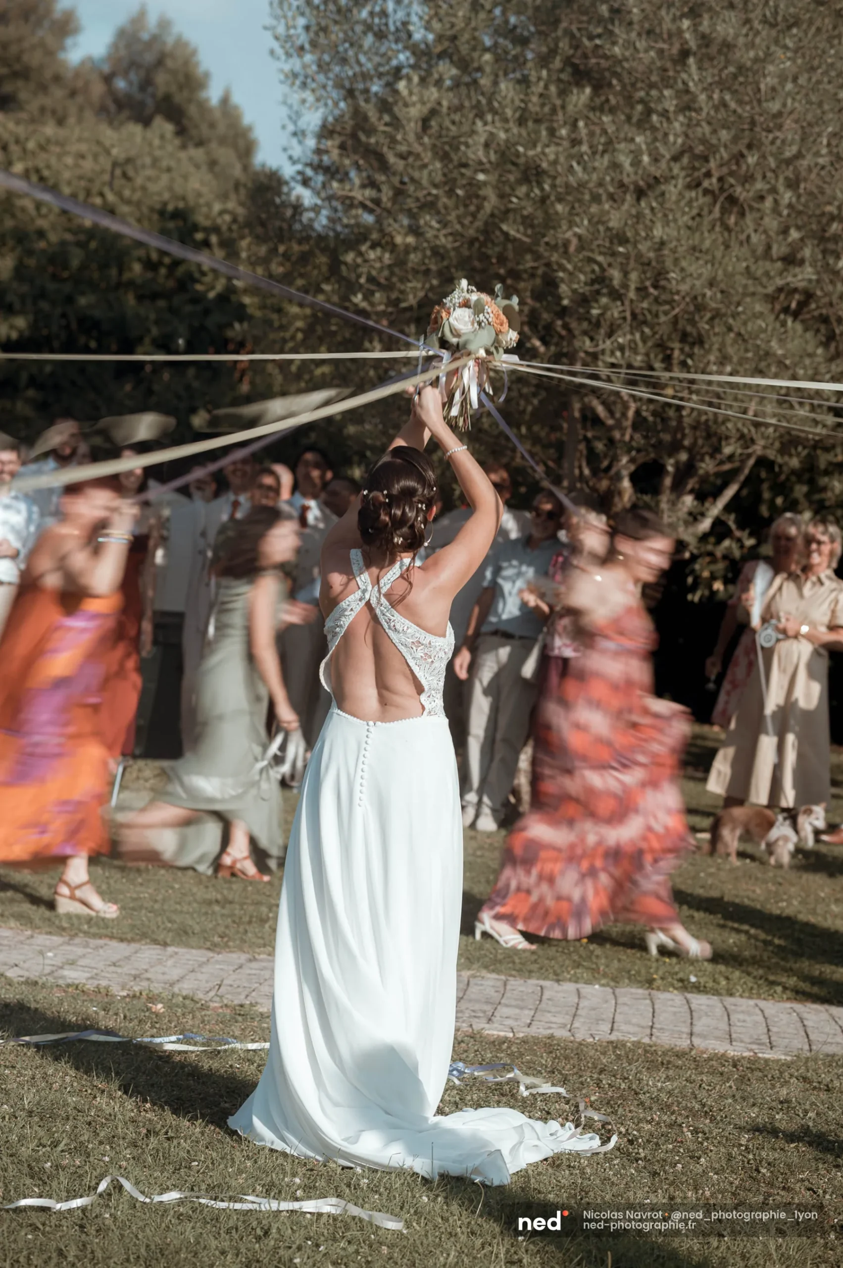 La mariée lève son bouquet pendant le jeu des rubans, les invitées en mouvement autour d'elle, Domaine de Bellevue