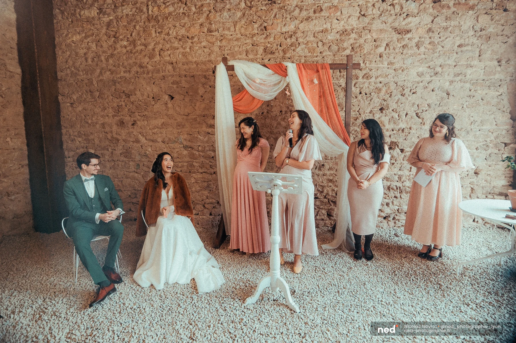Mariés assis riant pendant discours cérémonie laïque mur pierre arc tissu, mariage au Domaine de Tourieux