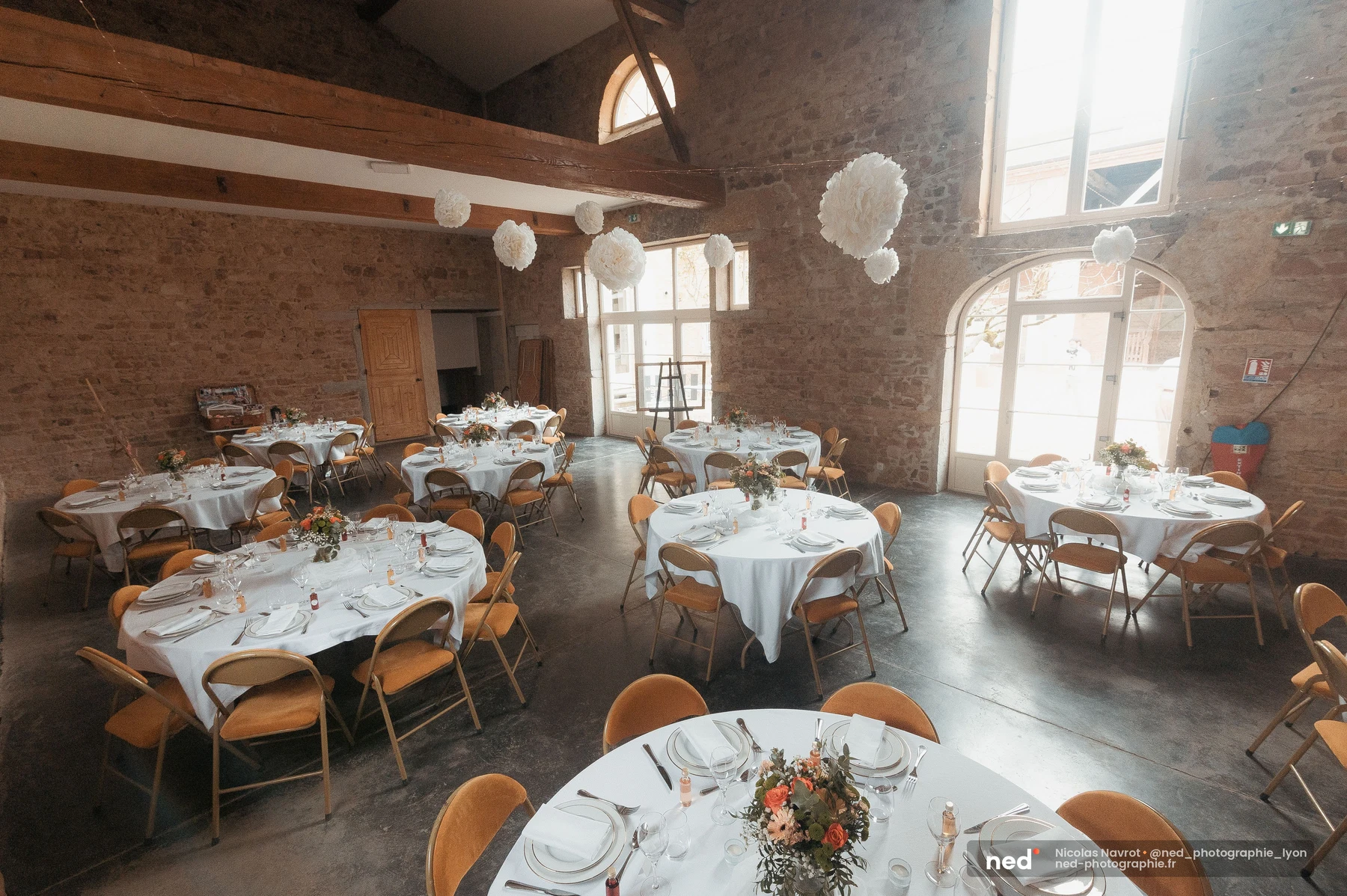 Salle de réception mariage en pierres apparentes avec tables rondes fleuries et pompons blancs, mariage au Domaine de To