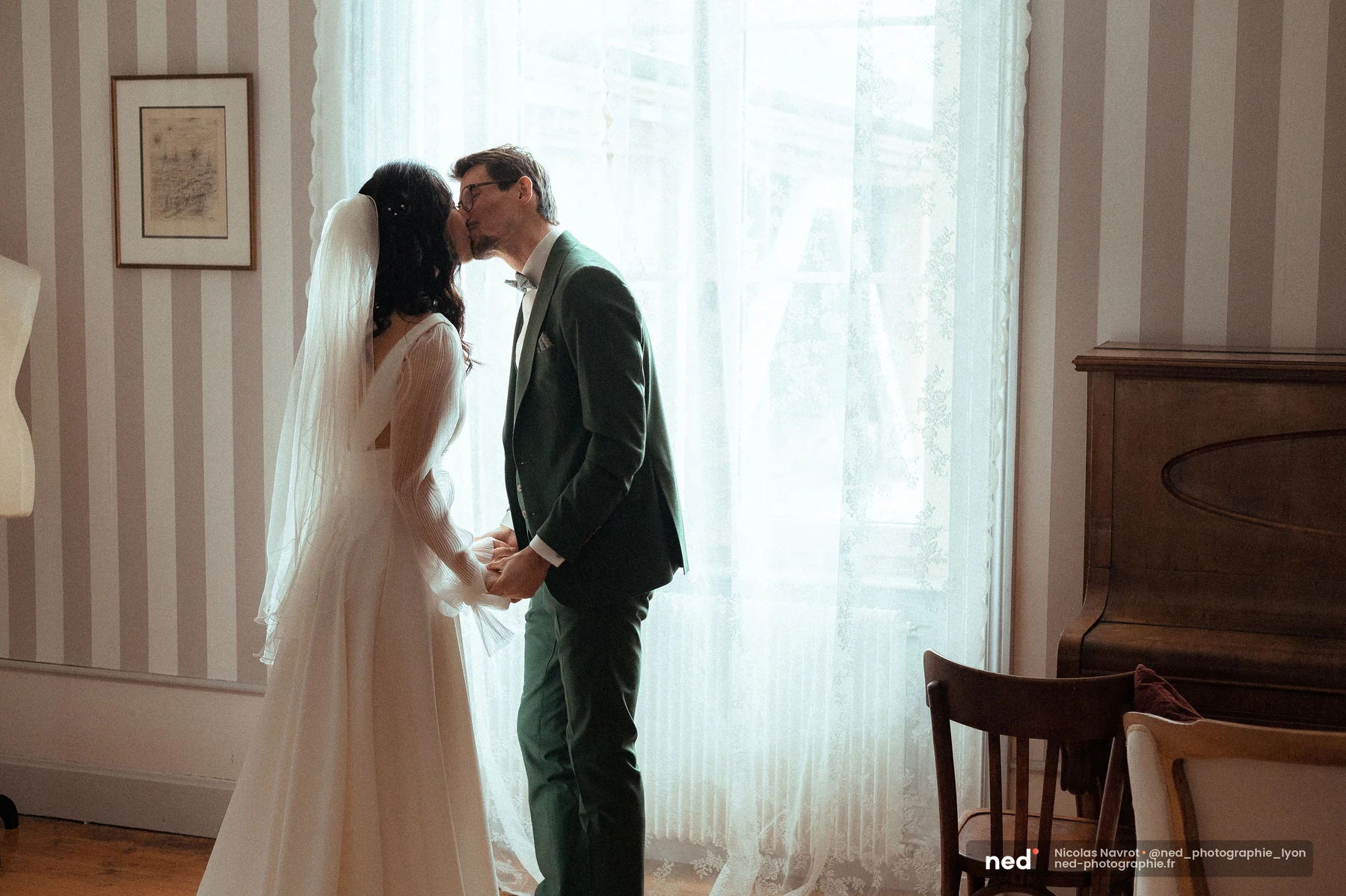 Mariés s'embrassant mains jointes contre-jour fenêtre voilages blancs intérieur, mariage au Domaine de Tourieux