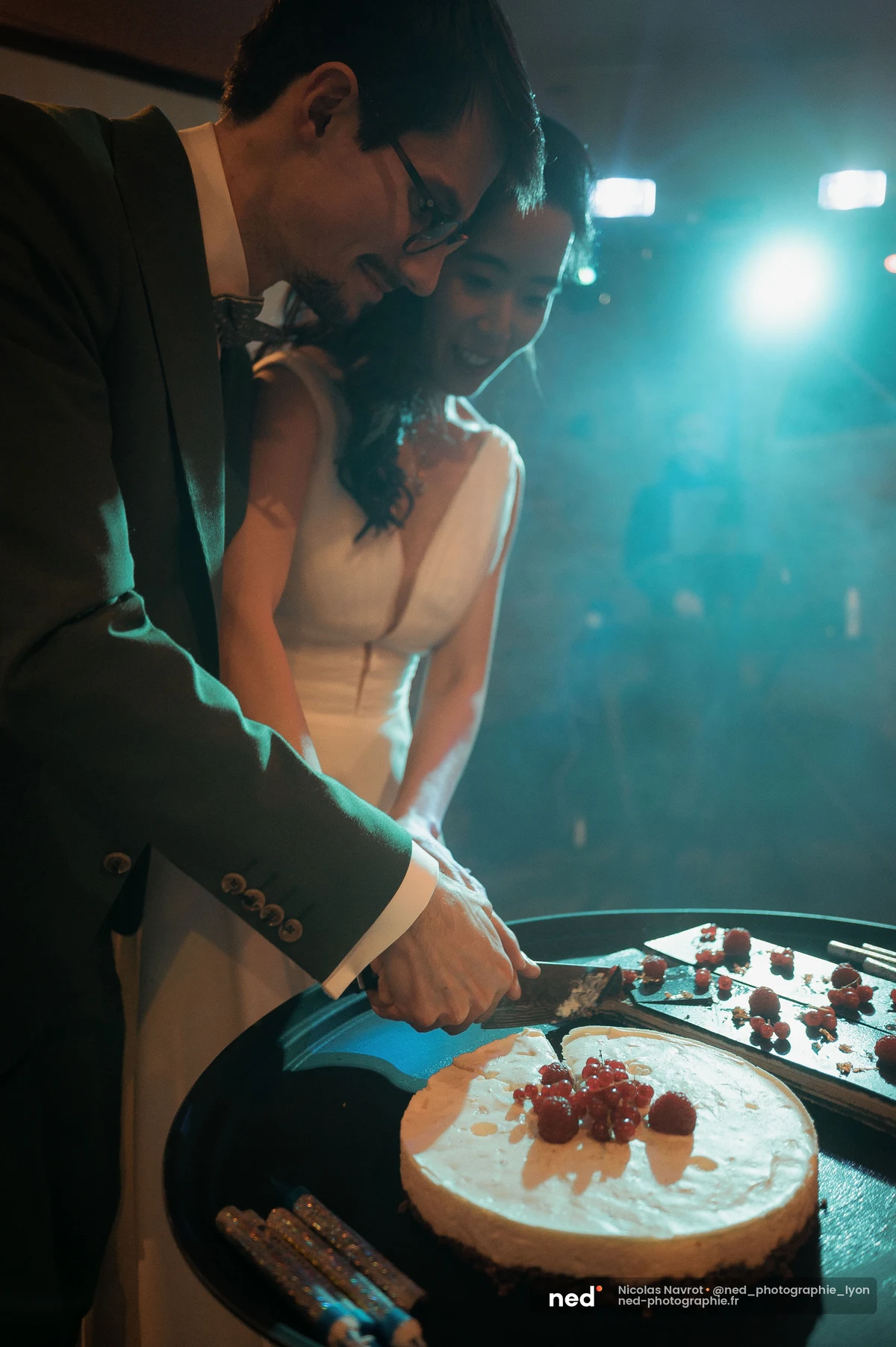 Mariés découpant ensemble entremets framboises sous lumière bleue soirée, mariage au Domaine de Tourieux
