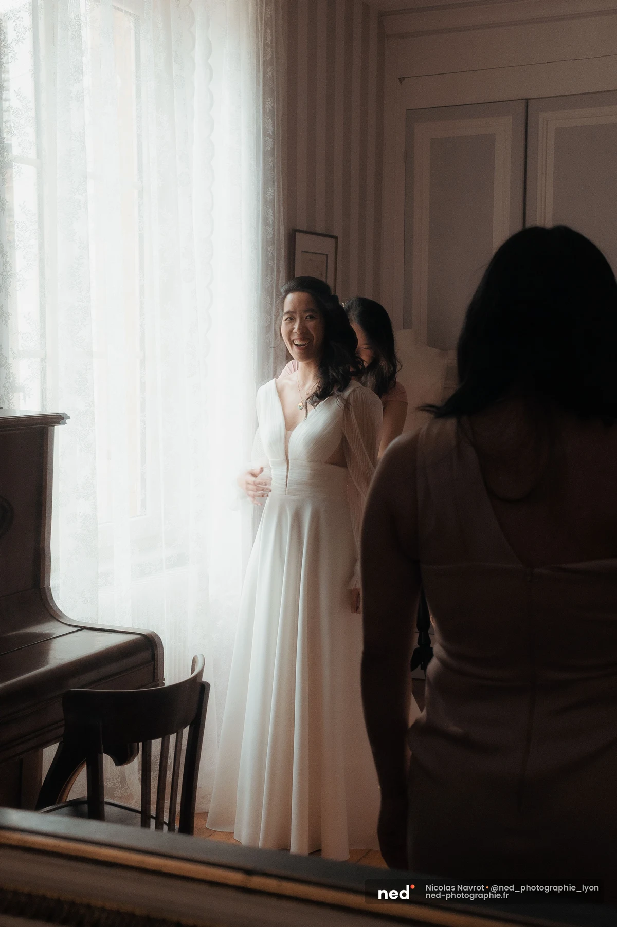 Mariée robe blanche fluide tulle devant fenêtre dentelle entourée de proches préparatifs, mariage au Domaine de Tourieux