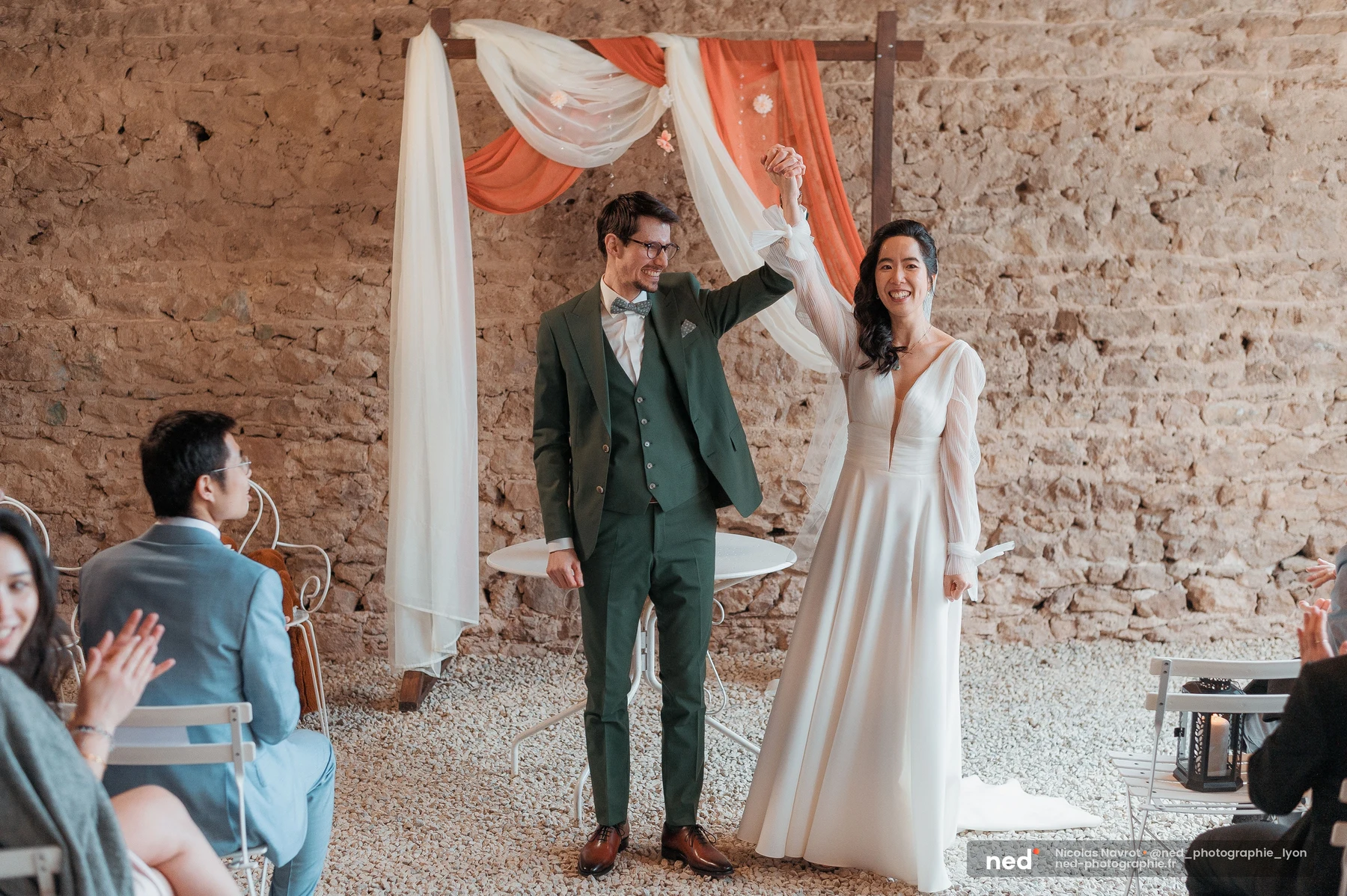 Mariés main levée sous arche de tissu blanc et terracotta, mur en pierres apparentes, mariage au Domaine de Tourieux
