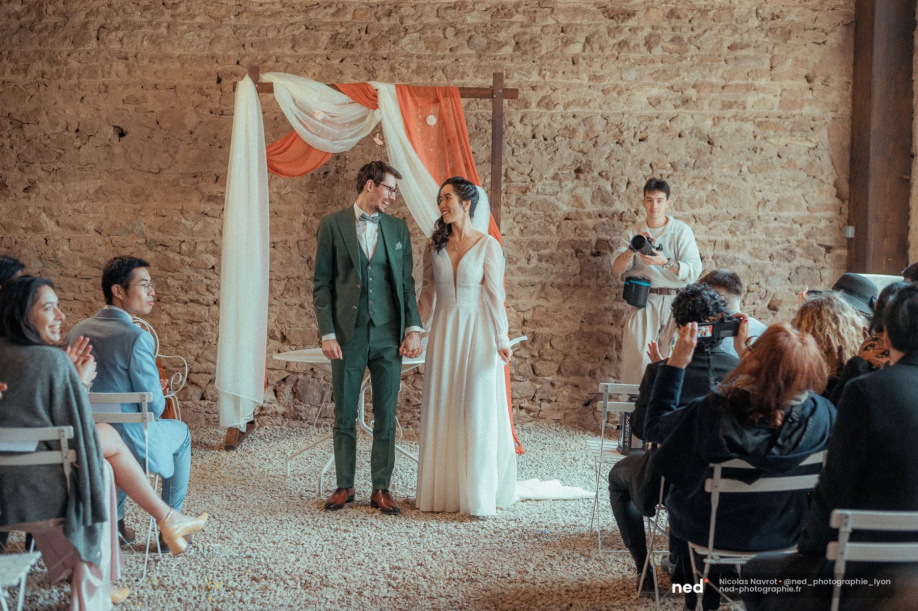 Mariés se regardant main dans la main devant une arche de tissu en grange en pierre, mariage au Domaine de Tourieux