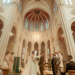 Mariée en robe blanche face au marié dans une église aux voûtes ogivales, famille et invités alignés de chaque côté, lumière naturelle des vitraux