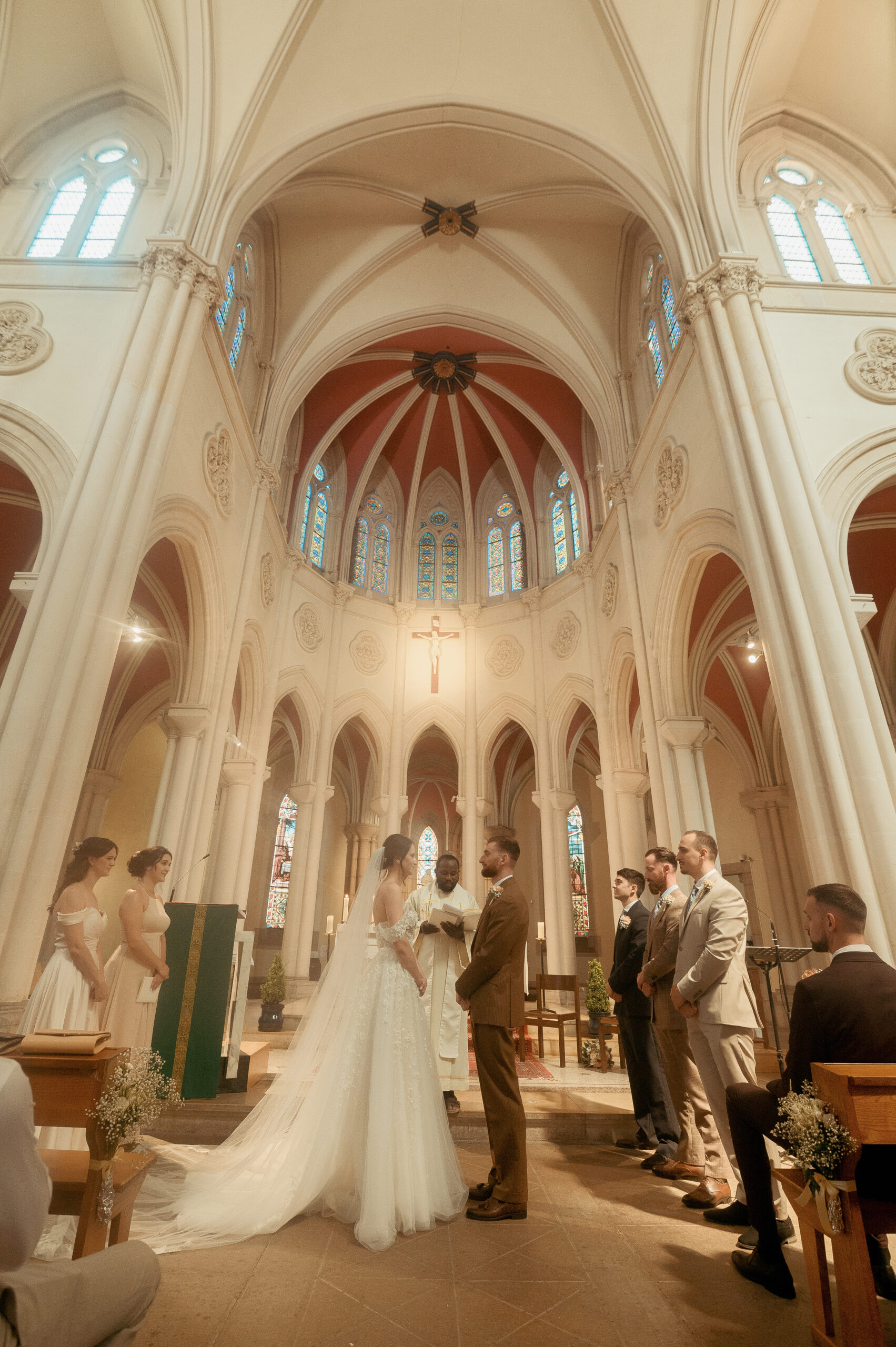 Mariée en robe blanche face au marié dans une église aux voûtes ogivales, famille et invités alignés de chaque côté, lumière naturelle des vitraux
