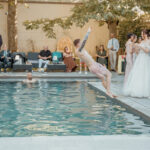 Marié en costume clair se jette dans une piscine intérieure pendant la réception, mariée en robe blanche regarde, invités assis autour