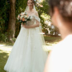 Mariée en robe blanche avec voile tient un bouquet de roses pêche et blanches sous un grand saule, son futur époux flou au premier plan