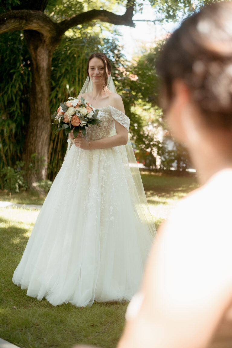 Mariée en robe blanche avec voile tient un bouquet de roses pêche et blanches sous un grand saule, son futur époux flou au premier plan