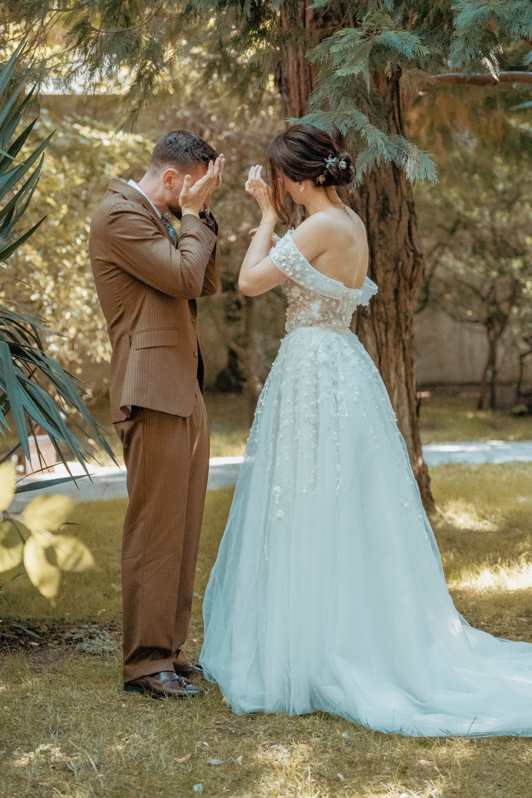 Mariee en robe blanche et marie en costume marron se regardent les mains sur le visage dans un jardin arbore